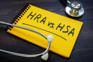 HRA vs. HSA is written on a yellow notebook alongside a stethoscope