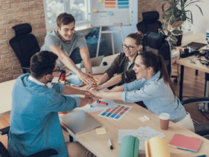 Group of young professionals in a modern office celebrating teamwork with a hands-in gesture around a shared desk filled with charts, laptops, and coffee.