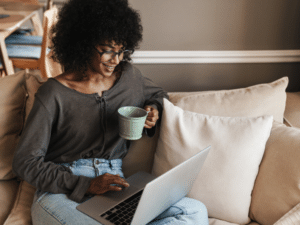 Woman sitting on a beige couch, smiling while using a laptop and holding a green mug, representing remote work or digital engagement.
